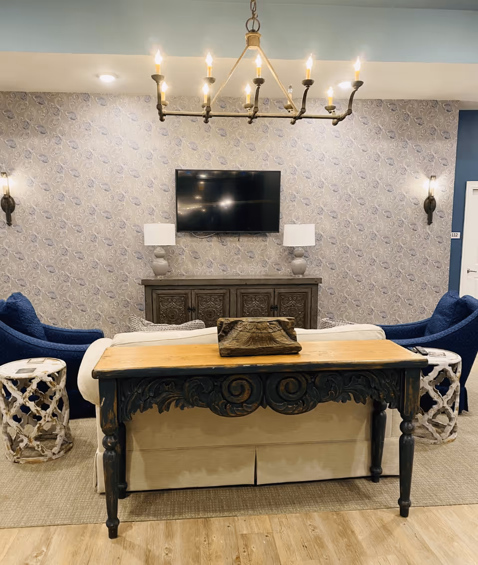 Lounge with a wooden console table behind a sofa, blue armchairs, patterned wallpaper, a wall-mounted TV, and a chandelier.