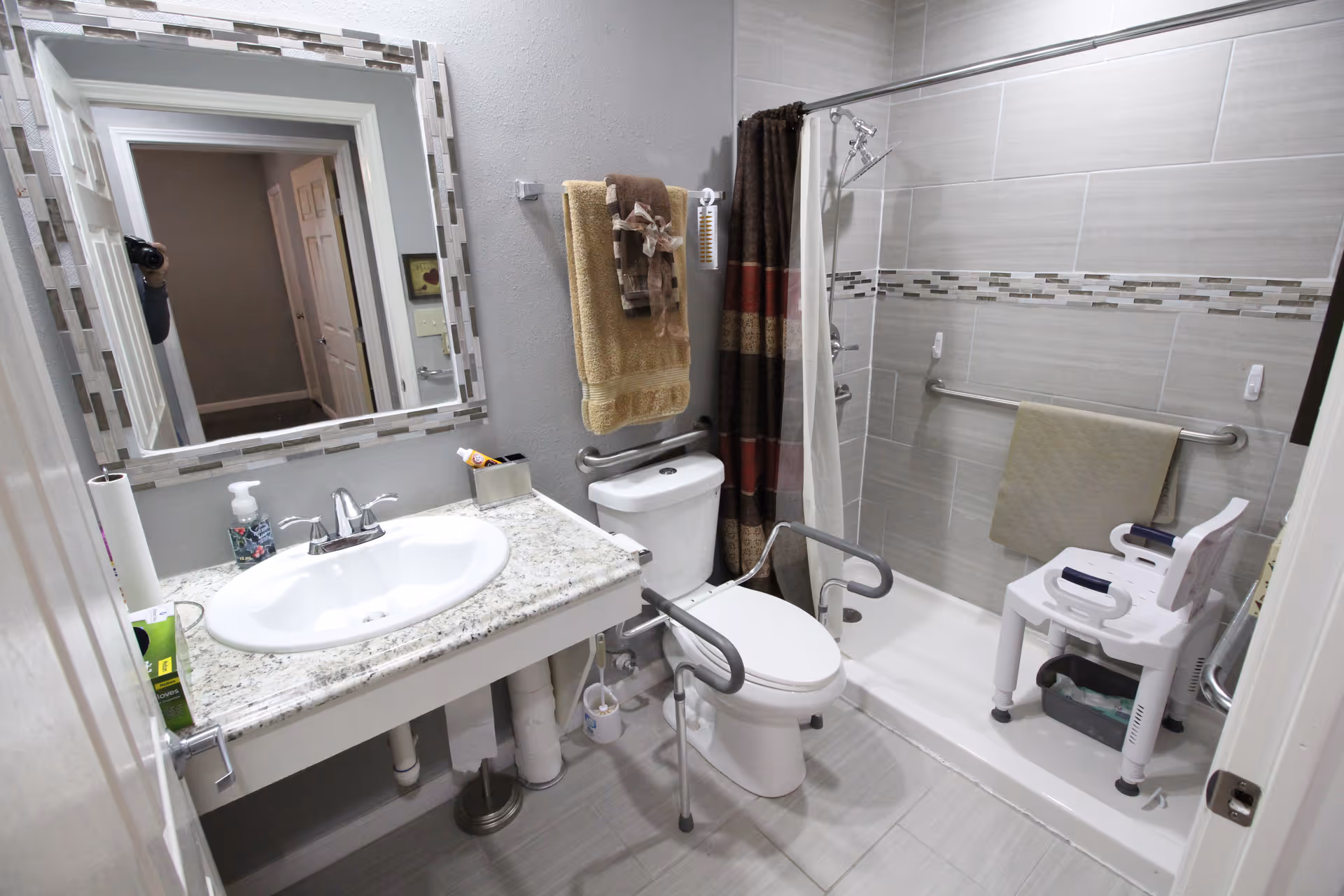 Accessible bathroom featuring a sink and mirror, a toilet with grab bars, and a roll-in shower with a shower chair.