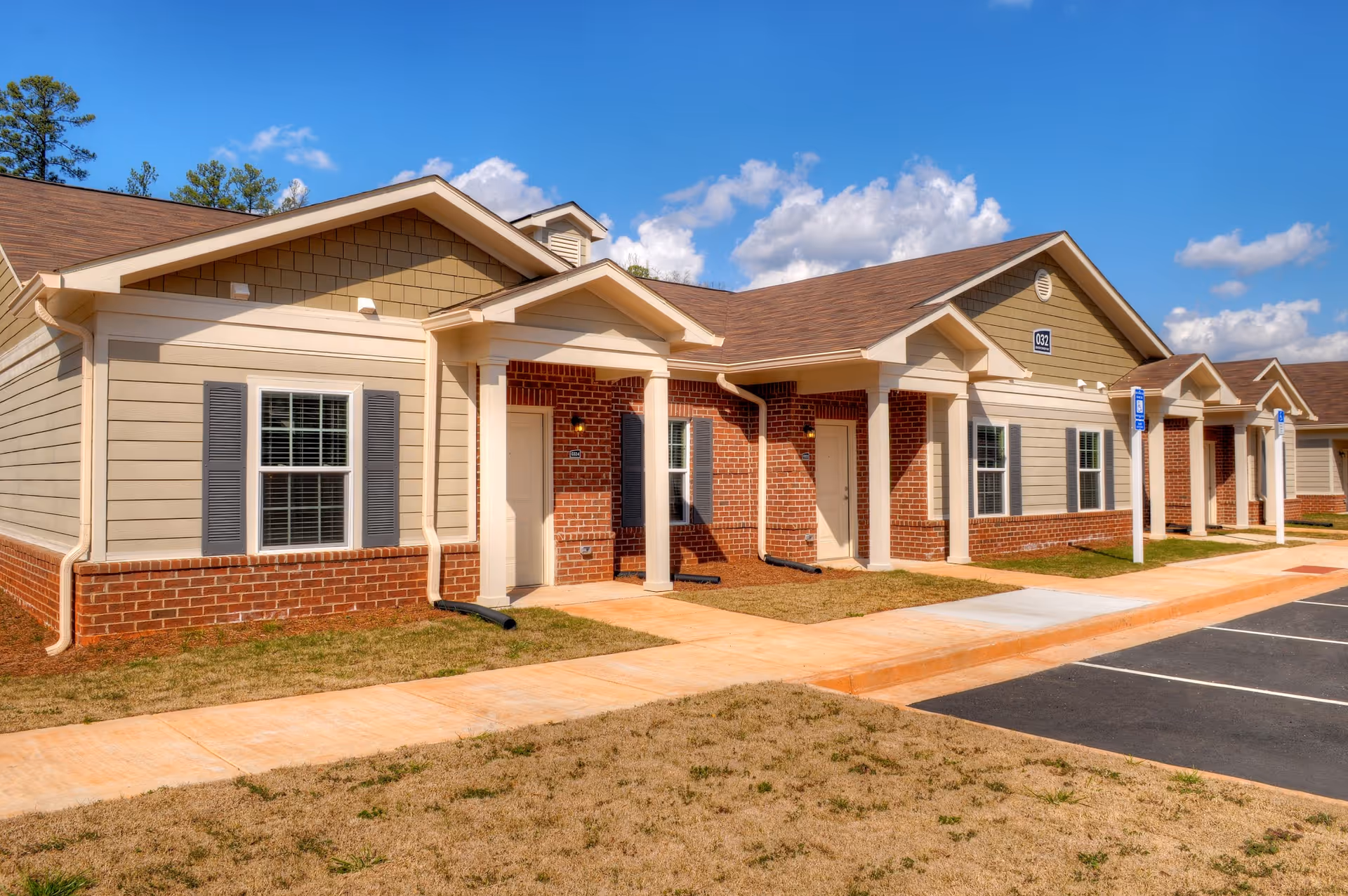 Exterior view of a single-story senior living facility building with beige siding, red brick accents, multiple windows with shutters, and several white entrance doors under small covered porches. The building is adjacent to a paved parking area with marked handicapped parking spaces. The sky is clear with some clouds.