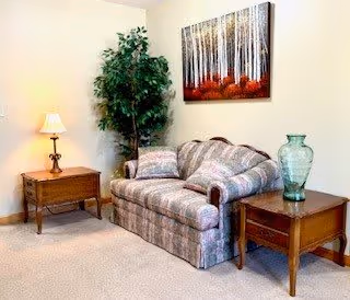 A cozy living room area with a patterned loveseat sofa flanked by two wooden side tables. One side table has a lamp with a beige shade, and the other has a large green glass vase. A tall artificial plant is placed in the corner behind the sofa, and a framed artwork depicting a forest with white birch trees and red foliage hangs on the wall above the sofa.