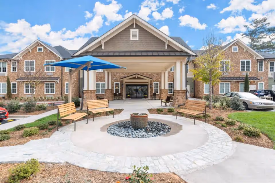 Front entrance of a brick senior living building with a covered porte-cochère, benches, a circular fountain, and landscaping.