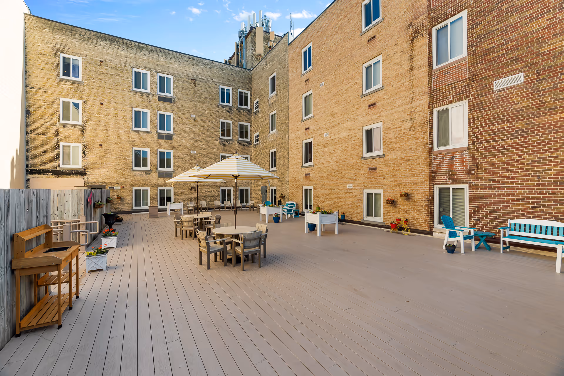 A spacious outdoor patio area with wooden decking, several round tables with chairs, and large umbrellas providing shade. The patio is surrounded by a multi-story brick building with multiple windows. There are also some potted plants, a wooden bench, and a small wooden shelving unit along the fence.