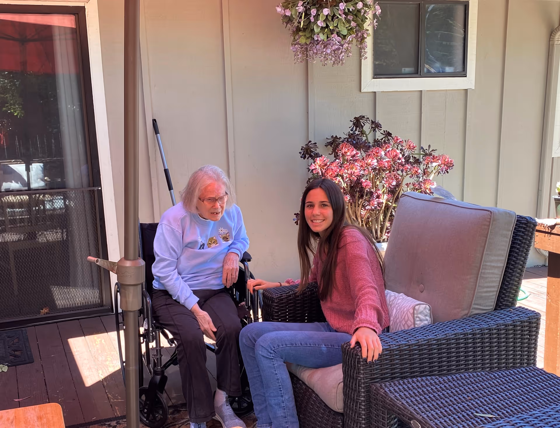 An elderly woman in a wheelchair and a younger woman sitting on a cushioned wicker chair are outside on a wooden deck. There are plants and flowers in the background, and a sliding glass door is visible to the left.