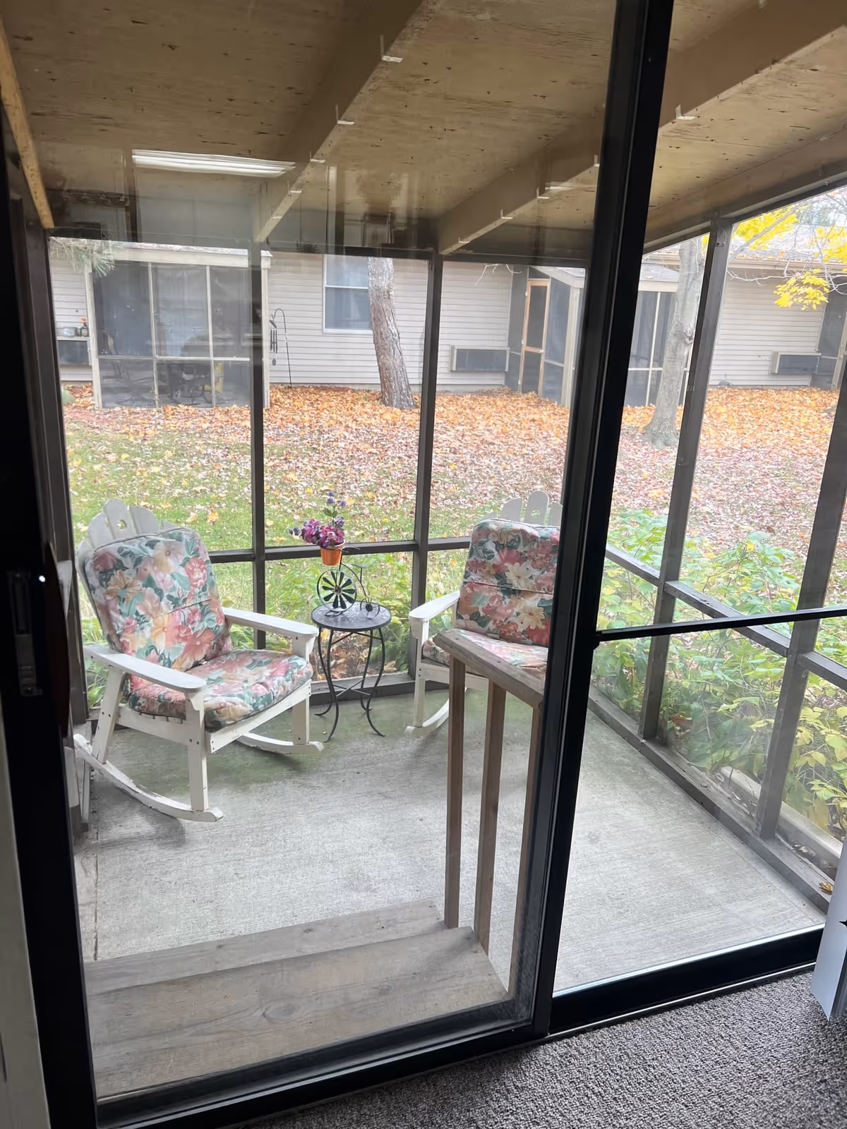 View through a sliding glass door of a screened-in porch with two white wooden rocking chairs with floral cushions and a small round metal table holding a small potted plant. Outside the porch, there is a grassy area with fallen autumn leaves and trees, and a building with windows and air conditioning units is visible in the background.