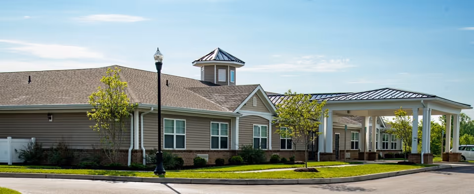 Exterior view of a single-story assisted living facility building with beige siding, multiple windows, a covered entrance supported by white columns, and a small tower-like structure on the roof. The building is surrounded by a well-maintained lawn, small trees, and a streetlamp under a clear blue sky.