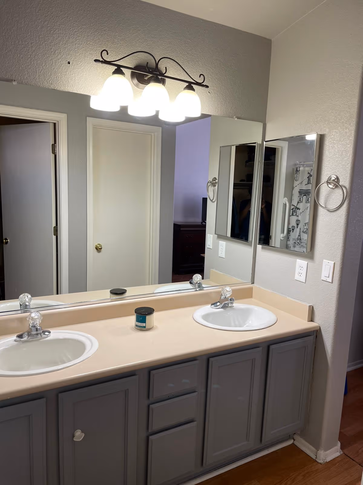 Bathroom vanity with two sinks, a large mirror, and a light fixture with four bulbs above. There is a candle placed on the countertop. Two doors are visible in the reflection of the mirror, and a towel ring is mounted on the wall next to the mirror.