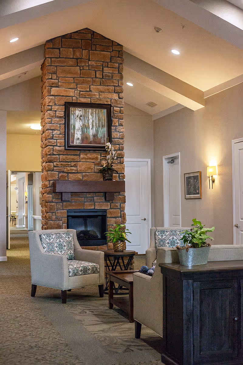 A cozy interior common area with a stone fireplace as the focal point. Two upholstered armchairs with patterned cushions face each other across a wooden coffee table. There are potted plants on the tables and a framed painting above the fireplace. The room has beige walls, carpeted floors, and soft lighting from wall sconces and recessed ceiling lights.