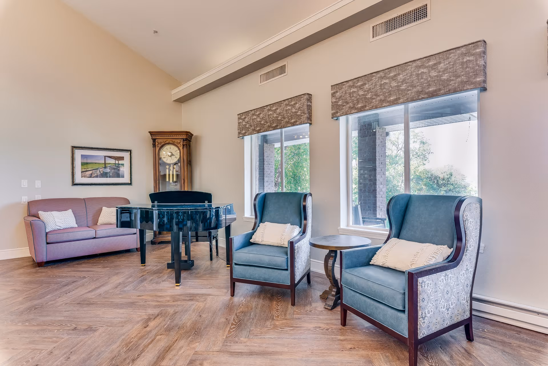 A bright and spacious living room area with two blue upholstered armchairs with white pillows, a small round wooden side table between them, a black grand piano, a pink sofa with two white pillows, a tall wooden grandfather clock, and large windows with gray valances letting in natural light.