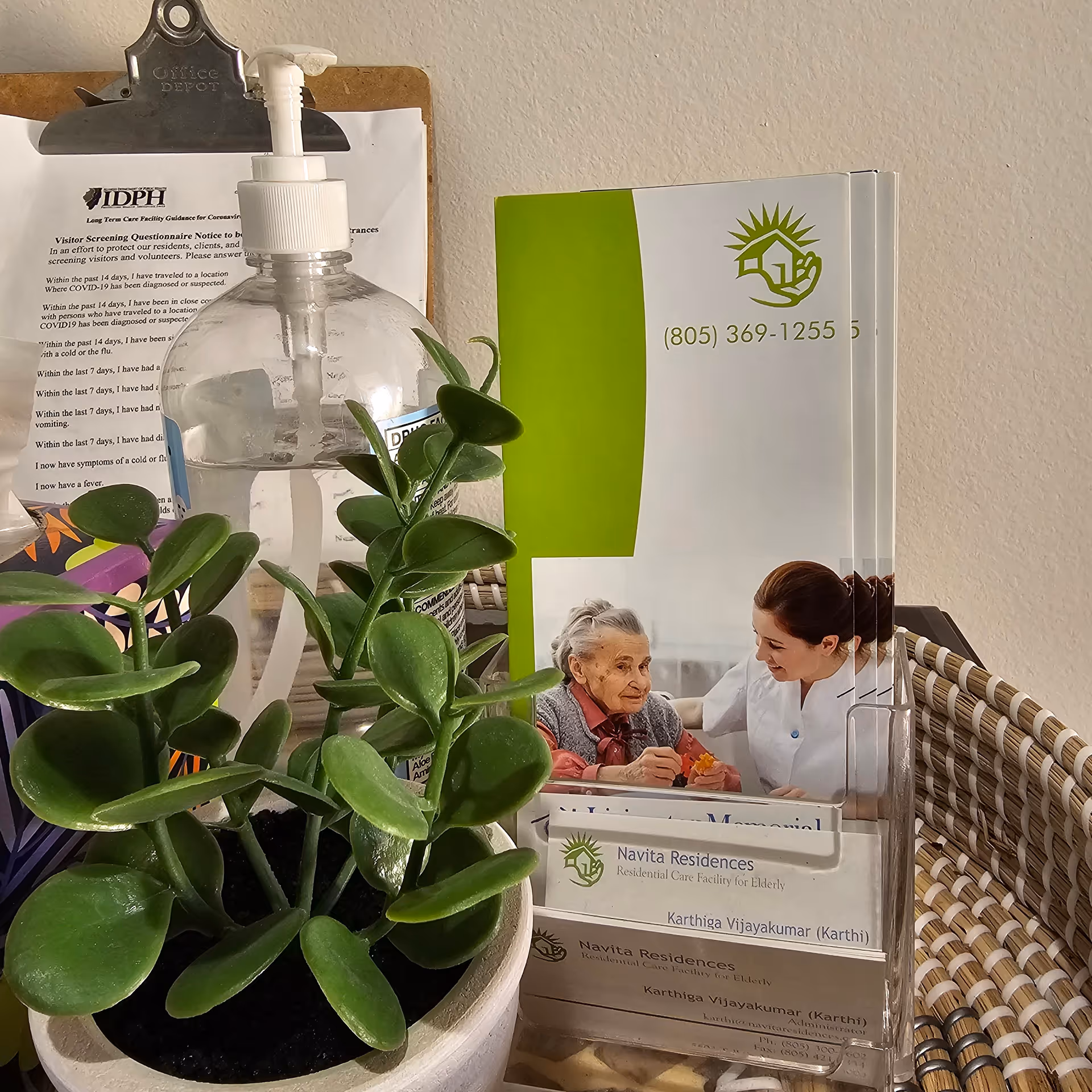 A small potted green plant, a clear bottle of hand sanitizer with a pump, and a brochure holder containing brochures for Navita Residences, a residential care facility for the elderly. Behind the items is a clipboard with a printed visitor screening questionnaire.