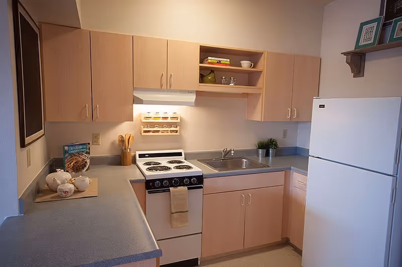 A compact kitchen with light wood cabinets, a white electric stove with four burners, a stainless steel sink, and a white refrigerator. The countertop has a tea set and a cookbook, and there are small decorative items on the shelves and counter. The kitchen has a clean and simple design.