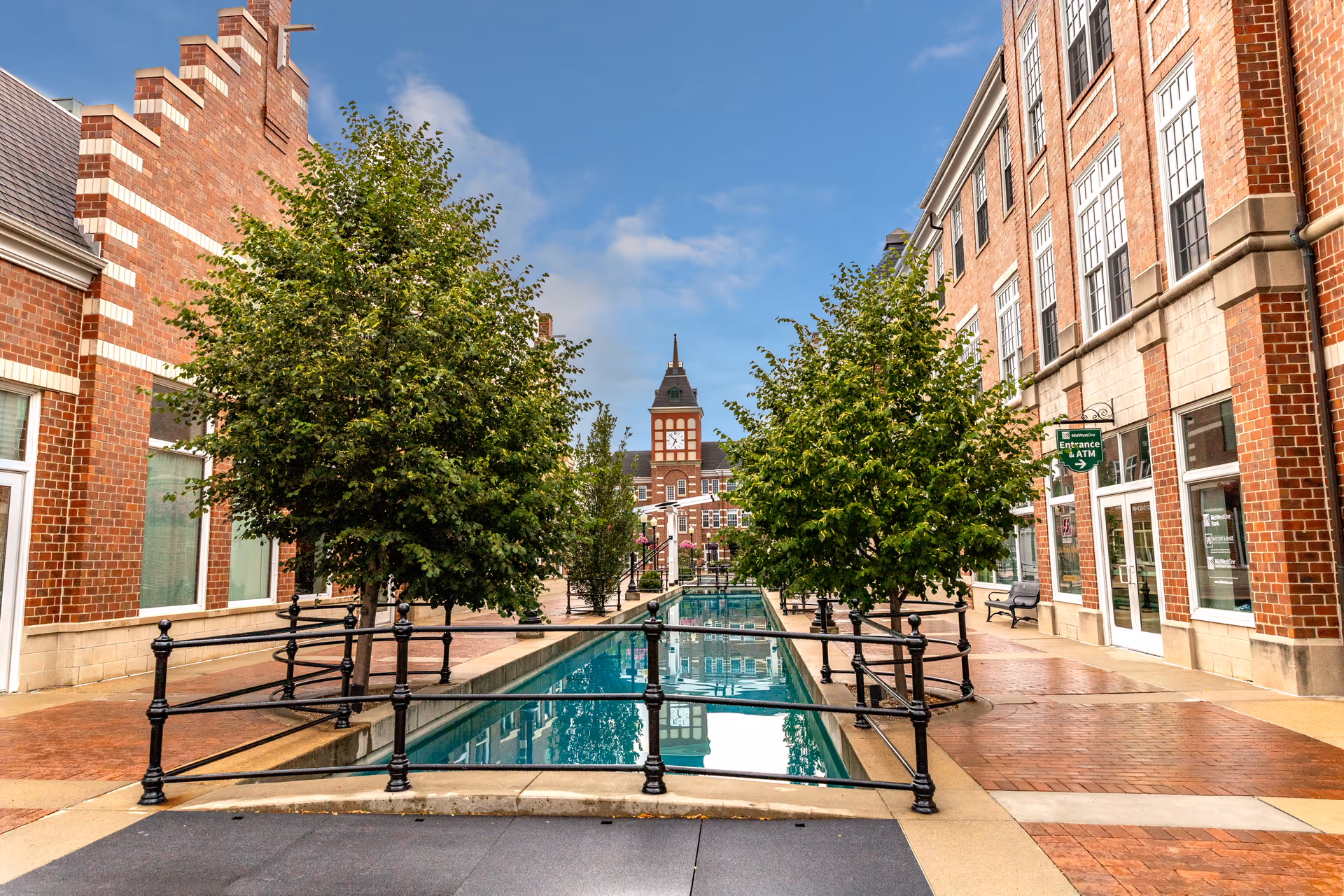 Outdoor view of a brick building complex with a narrow reflecting pool lined with trees and black railings in the center. The sky is blue with some clouds, and a clock tower is visible in the background.