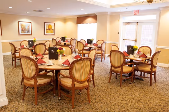 A well-lit dining room with several round wooden tables, each surrounded by four upholstered chairs with striped cushions. The tables are set with folded red napkins, cups, and small flower arrangements. The room has beige walls adorned with framed artwork and a carpeted floor. A glass exit door is visible in the background.