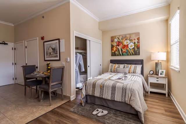 A cozy senior living studio apartment featuring a neatly made bed with patterned bedding and multiple pillows, a bedside table with a lamp, clock, and framed photo, a small dining table with two chairs, an open closet with robes hanging inside, and a window letting in natural light. The walls are painted beige and decorated with a colorful floral painting above the bed.