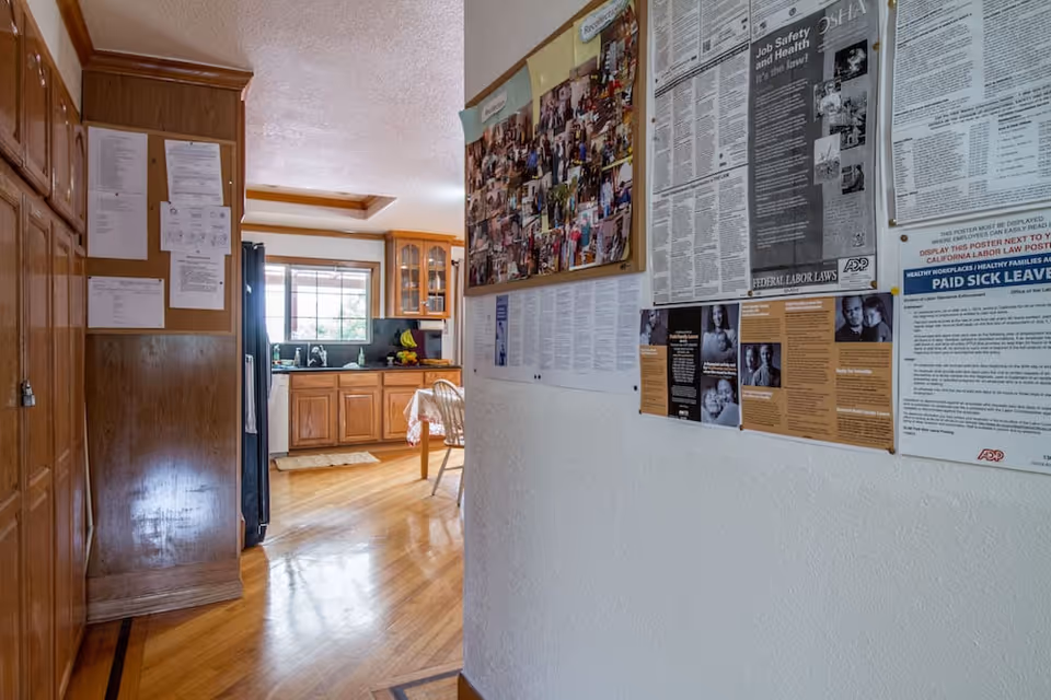 View down a hallway with wooden cabinets on the left and a bulletin board with various notices and photos on the right wall. At the end of the hallway, there is a kitchen area with wooden cabinets, a window, and a table with chairs.