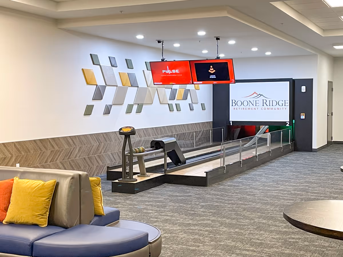 Indoor recreational area at Boone Ridge Retirement Community featuring a small bowling alley with two lanes, overhead screens, and modern wall art. There is a curved seating area with colorful cushions in the foreground.