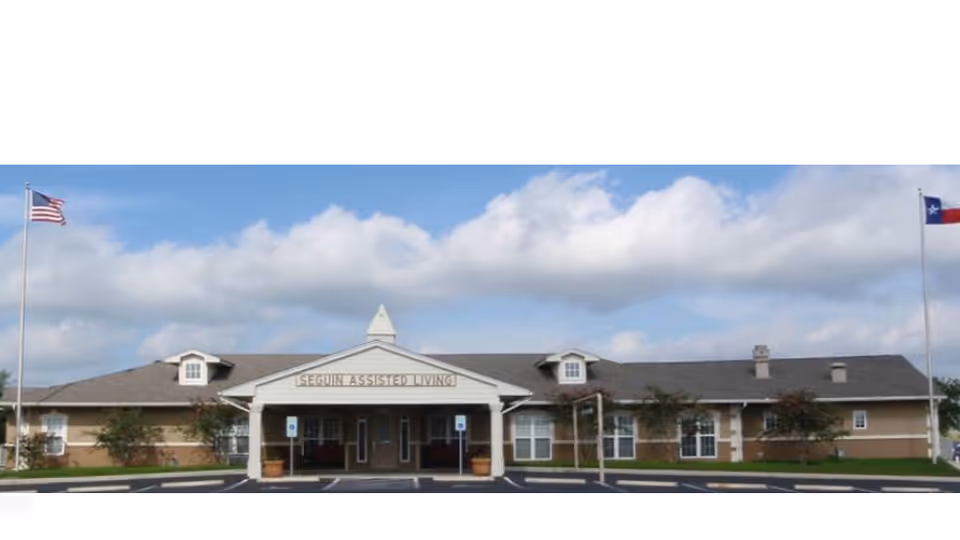 Front exterior view of Seguin Assisted Living facility with a covered entrance, two flagpoles displaying the American and Texas flags, and a clear blue sky with some clouds.