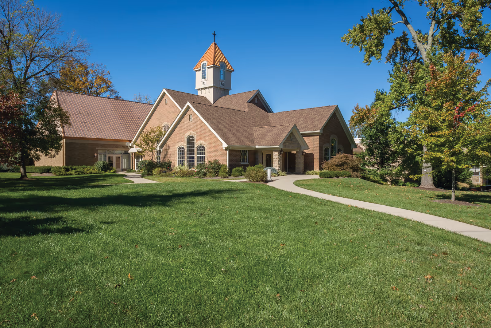 A brick building with a peaked roof and a small tower topped with a cross, surrounded by green grass and trees under a clear blue sky.