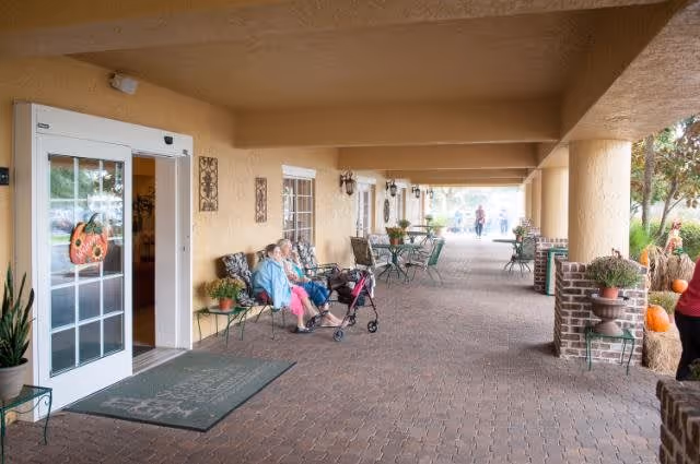 Covered outdoor walkway at Hunt Trace facility with two elderly women sitting on a bench, one using a walker. The walkway has brick flooring, beige walls, and several tables and chairs along the side. There are plants and seasonal decorations including pumpkins near the entrance door.