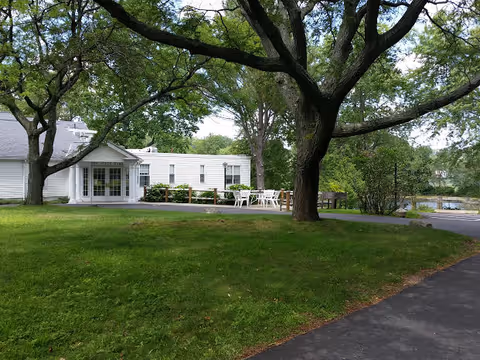 A white single-story building with large windows and a patio area with white outdoor chairs and tables, surrounded by green grass and large trees under a clear sky.