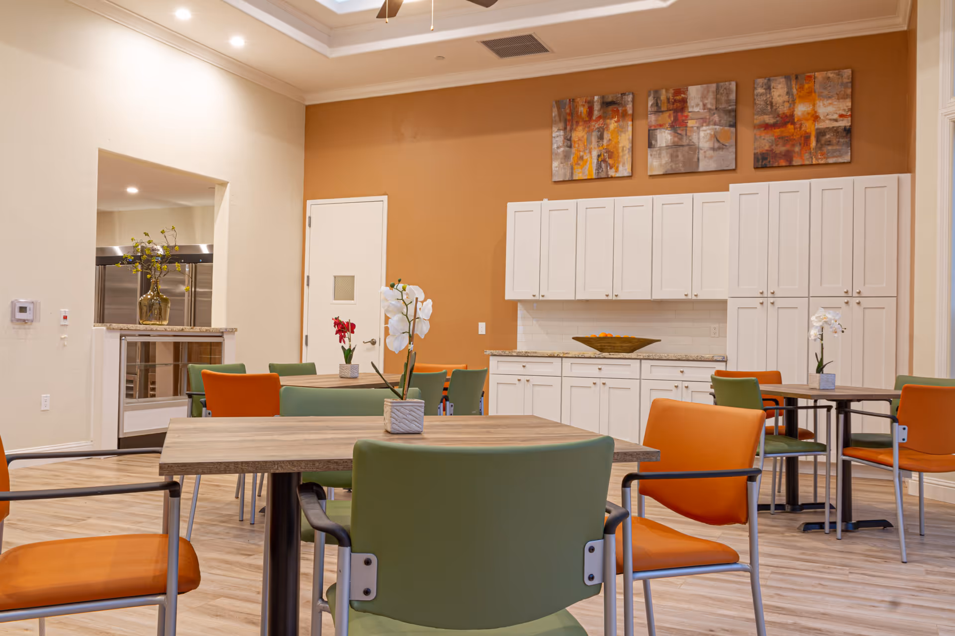 A bright dining room with wooden tables and chairs in green and orange upholstery. The room features white cabinets along a warm beige wall, with three abstract paintings hanging above. There are small flower arrangements on the tables and a large bowl on the countertop. The floor is light wood, and there is an open pass-through window to a kitchen area.