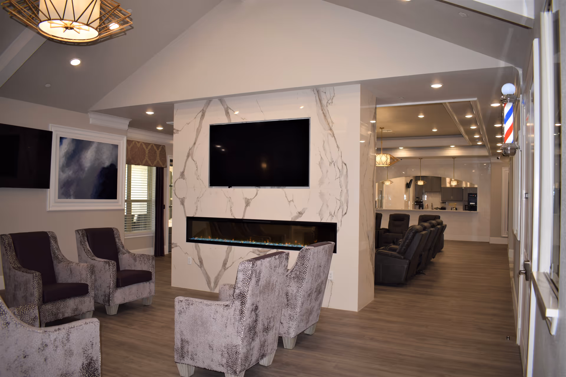 Interior view of a modern senior living facility lounge area with four upholstered armchairs arranged around a wall-mounted flat screen TV and a long electric fireplace set in a marble-patterned wall. In the background, there are additional recliner chairs and a kitchen area with pendant lighting. A barber pole is mounted on the right wall near the entrance.