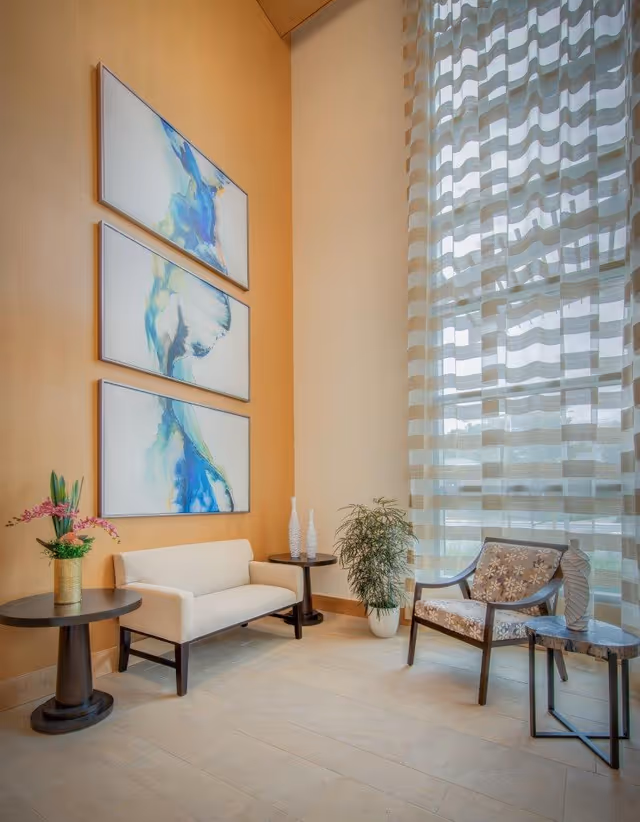 A bright and modern sitting area with a cream-colored loveseat, a floral-patterned armchair, two round side tables with decorative vases and a plant, and three abstract blue and white paintings on a wooden wall. Large windows with sheer striped curtains allow natural light to fill the space.