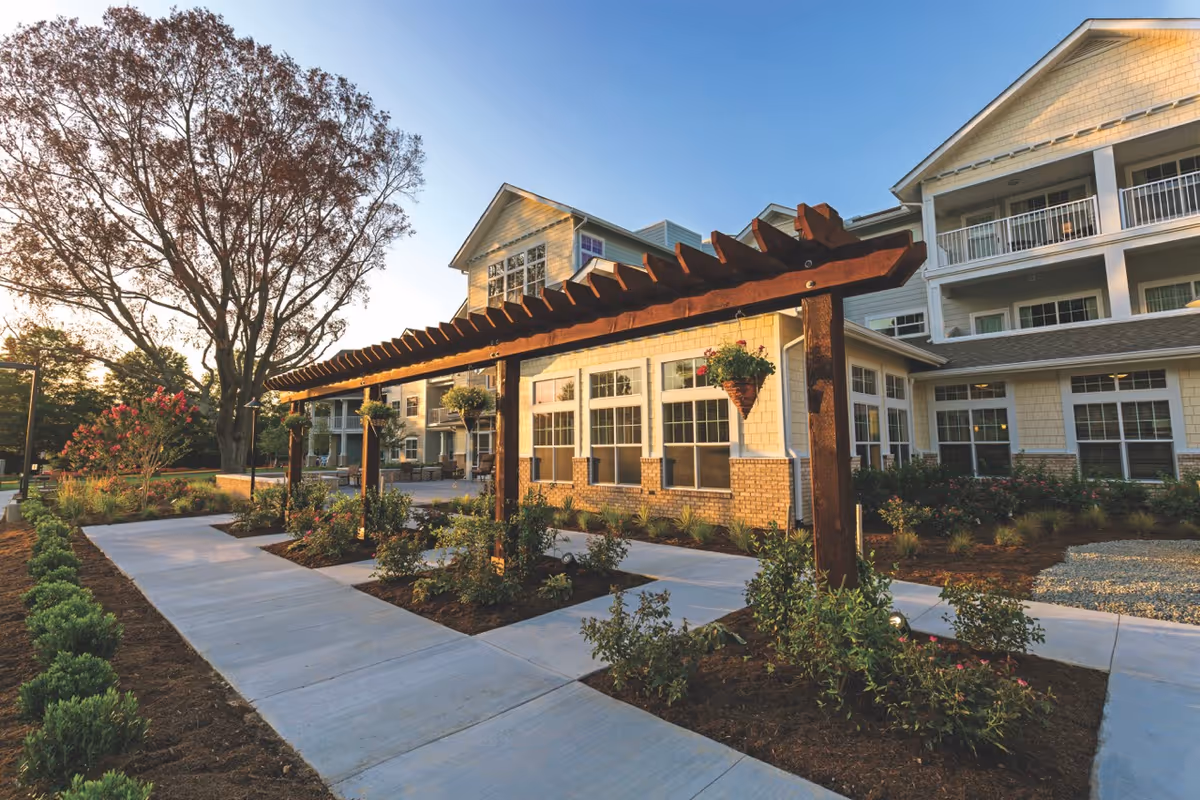 Front exterior of a multi-story senior living building with a wooden pergola, landscaped walkways, and hanging planters at sunset.