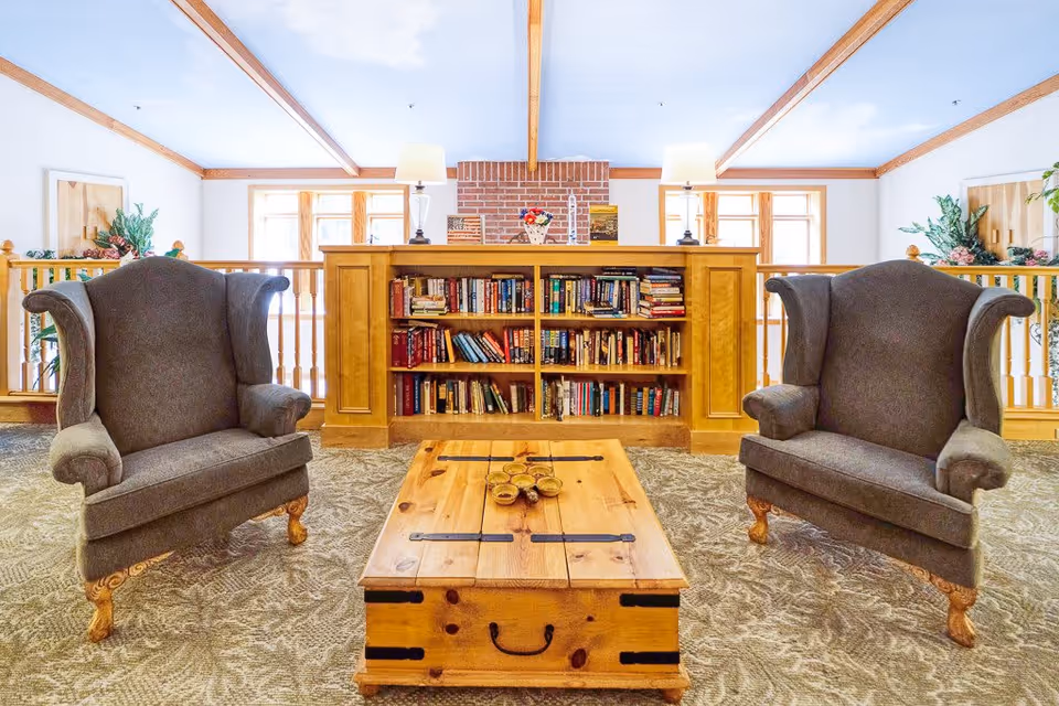 A cozy sitting area in a senior living facility with two gray upholstered armchairs facing a wooden coffee table. Behind the chairs is a wooden bookshelf filled with books, topped with two table lamps and decorative items. The room has a carpeted floor, wooden railing, and large windows letting in natural light.