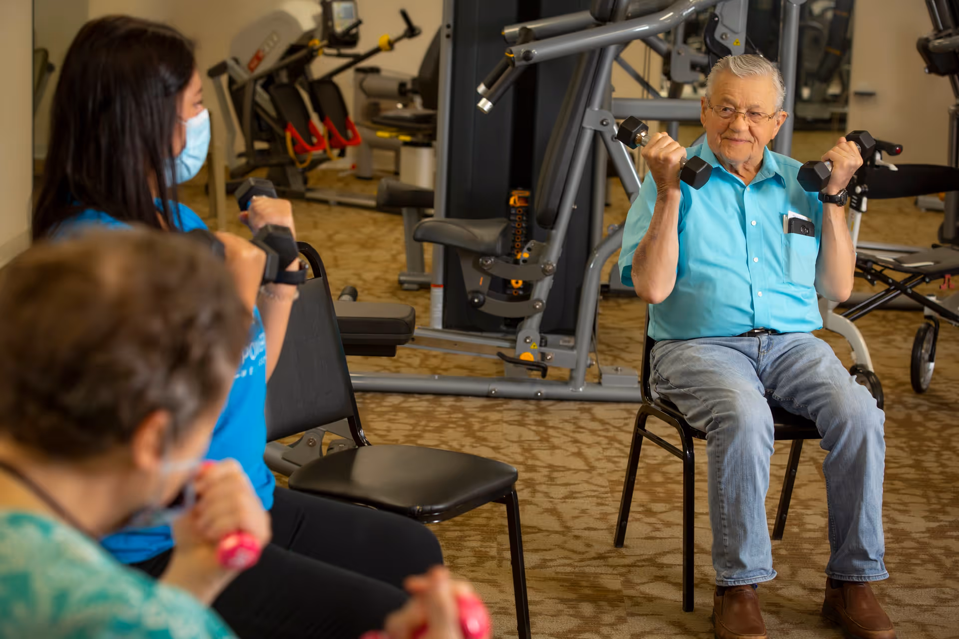 An elderly man sitting on a chair in a gym lifting dumbbells, accompanied by two other people also holding dumbbells, with exercise equipment visible in the background.