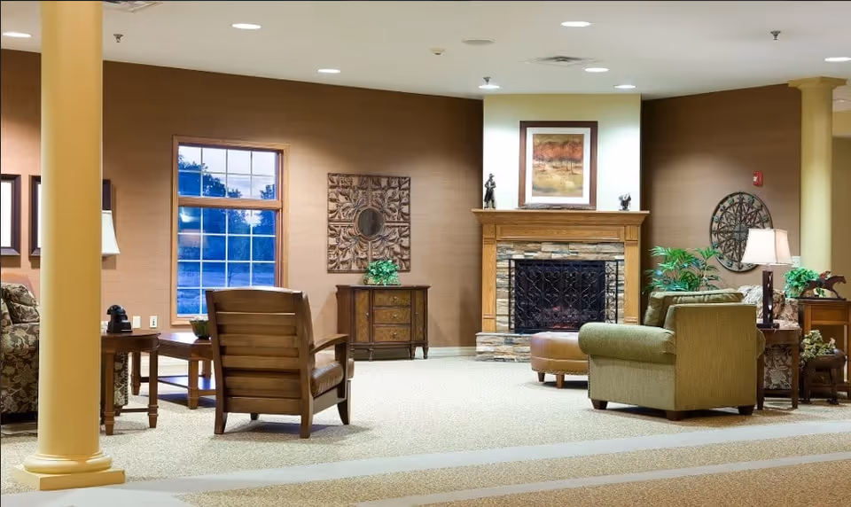 A cozy living room area in a senior living facility with a stone fireplace, framed artwork above the mantel, and comfortable seating including armchairs and a sofa. The room has warm brown walls, decorative wall art, indoor plants, and soft lighting from table lamps. Large windows allow natural light to enter the space.