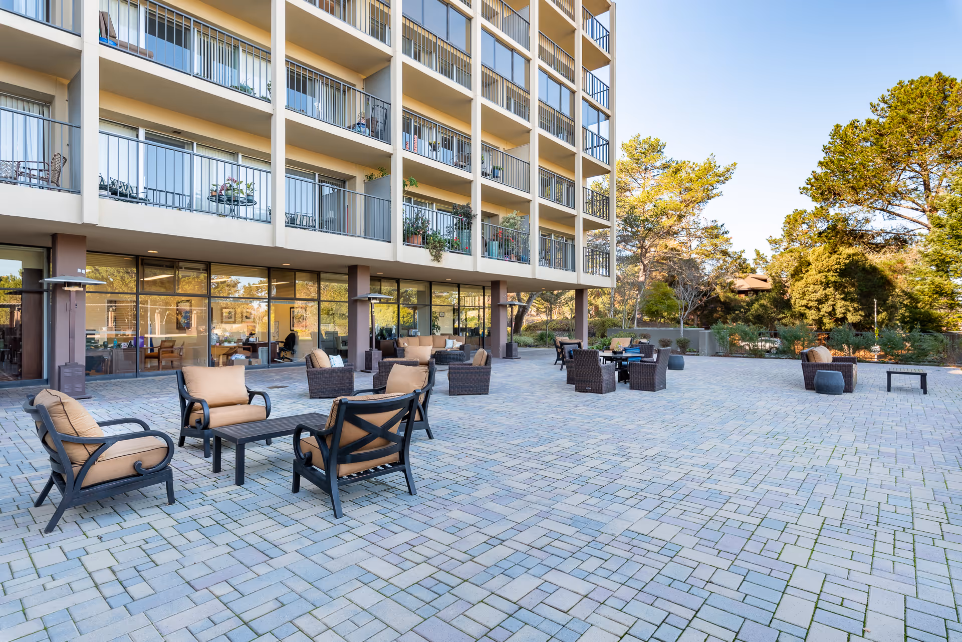 Outdoor patio area at The Park Lane with multiple seating arrangements including cushioned chairs and tables on a paved surface, adjacent to a multi-story building with balconies and large glass windows. Trees and greenery are visible in the background under a clear blue sky.