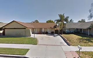 Front exterior view of a single-story residential building with a driveway, lawn, and palm trees under a clear sky.