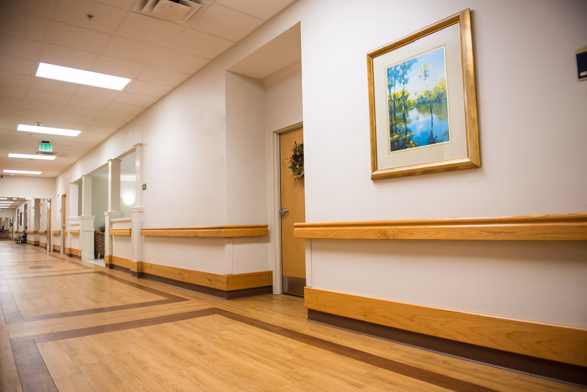Wide, well-lit interior hallway with wood handrails, framed artwork, and doors in a senior living facility.