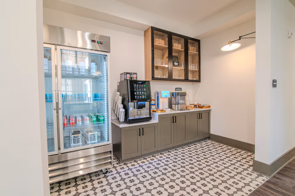A kitchenette area with a glass-door refrigerator, coffee and beverage machines on a countertop, upper cabinets, and patterned tile flooring.