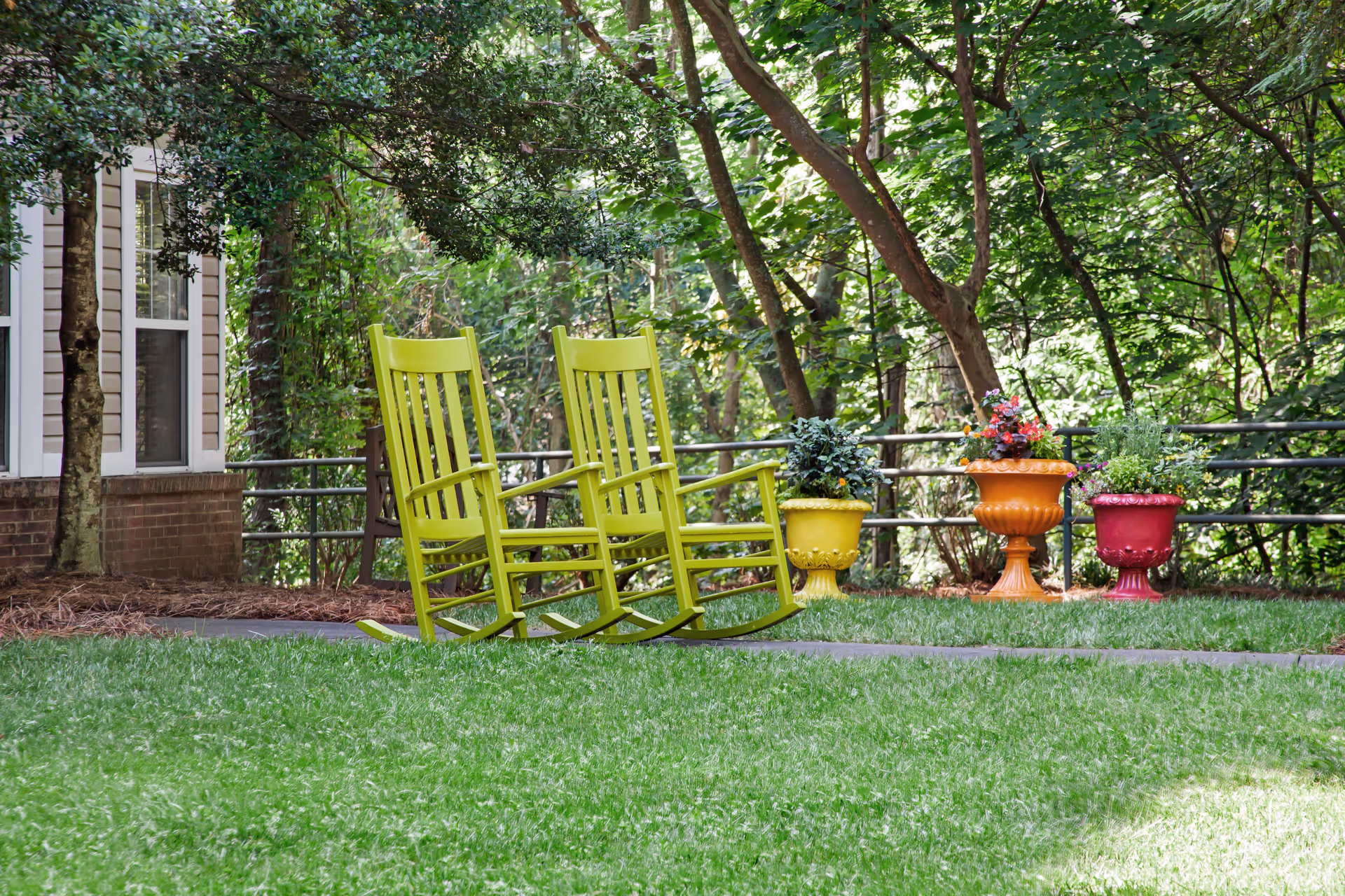 Two lime-green rocking chairs on a lawn beside colorful planters and trees outside a building.