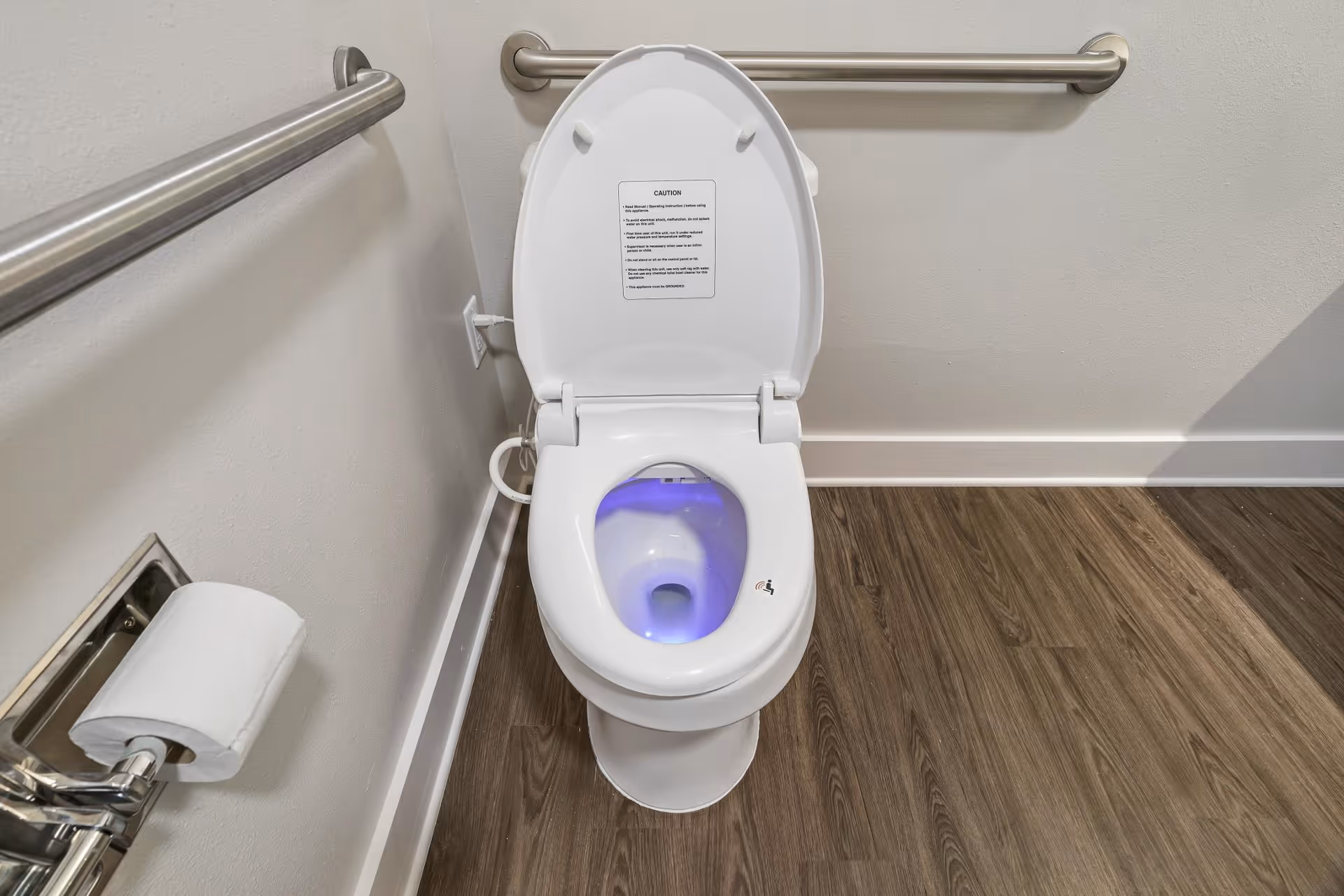 A white toilet with the lid open, showing a blue light inside the bowl. The toilet is installed in a bathroom with wood-look flooring, white walls, and stainless steel grab bars on the walls. A toilet paper holder with a roll of toilet paper is mounted on the left wall.