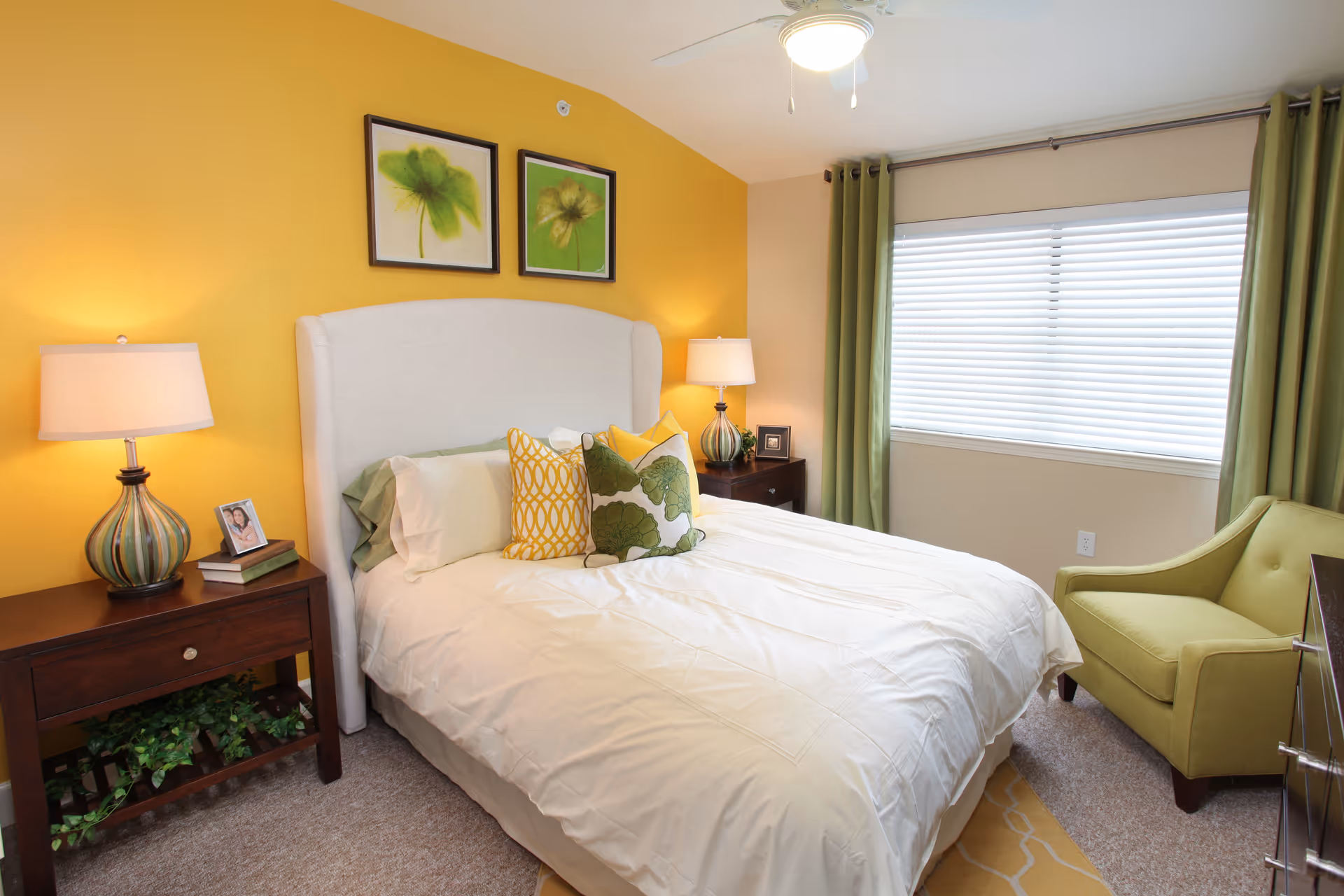 A cozy bedroom with a white upholstered bed featuring white and green pillows. The walls are painted yellow and beige, with two framed green floral artworks above the bed. There are two wooden nightstands with lamps on either side of the bed, a green armchair near a window with white blinds and green curtains, and a ceiling fan with a light.