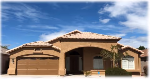 Exterior front view of a single-story house with a tiled roof, a two-car garage, and a small front yard with bushes and a tree under a partly cloudy blue sky.