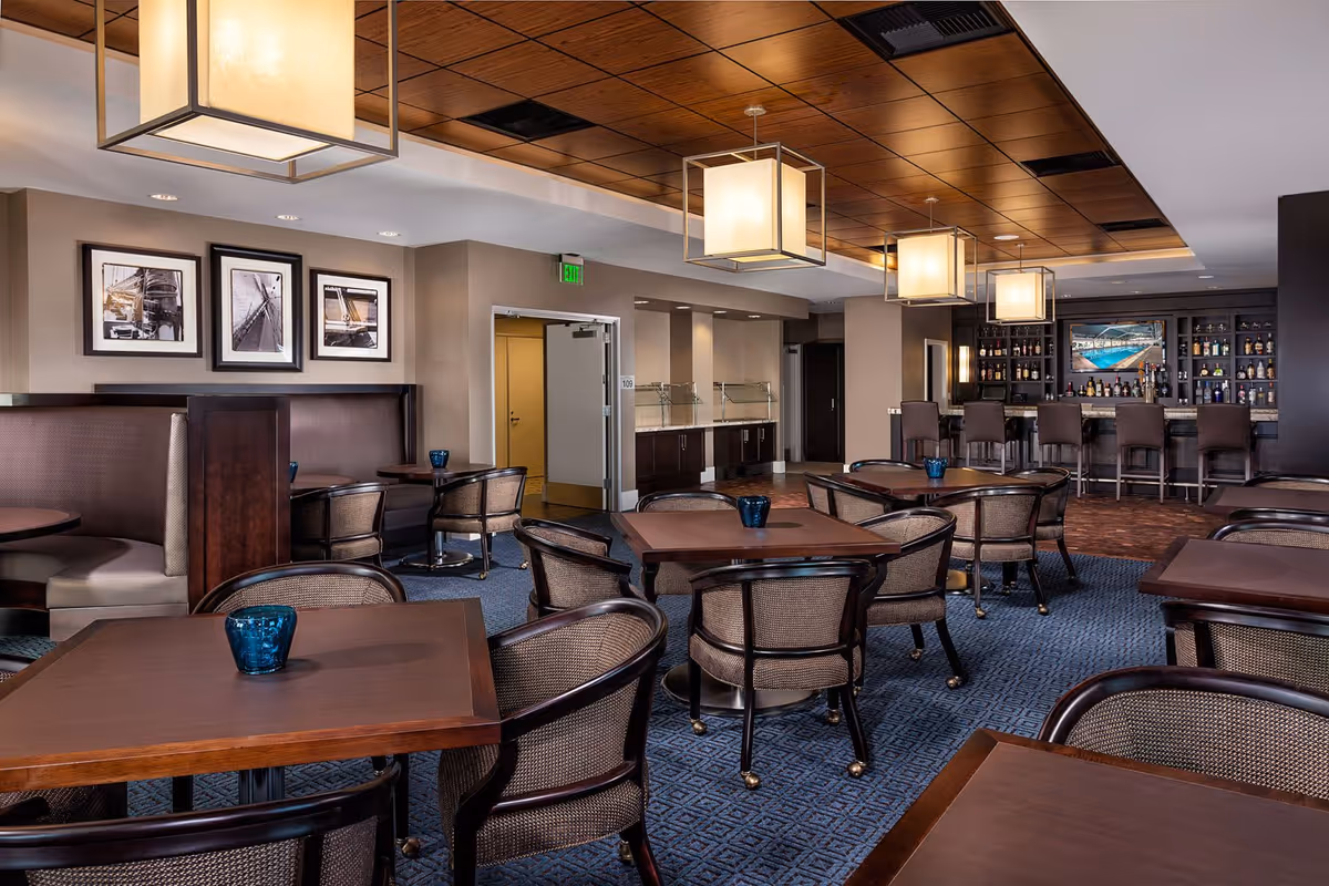 Interior view of a dining area with multiple wooden tables and cushioned chairs arranged neatly. The room features a blue carpet with a geometric pattern, a wooden ceiling with modern square pendant lights, and a bar area stocked with various bottles and a TV screen. There are framed black and white pictures on the wall and a doorway leading to another room.