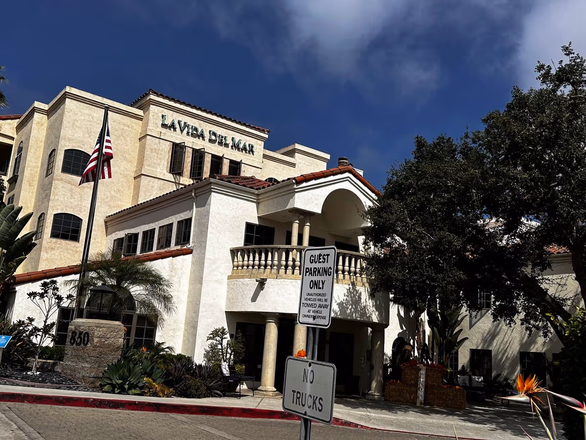 Front exterior of the La Vida Del Mar building with an arched entrance, American flag, landscaping, and guest parking signs.
