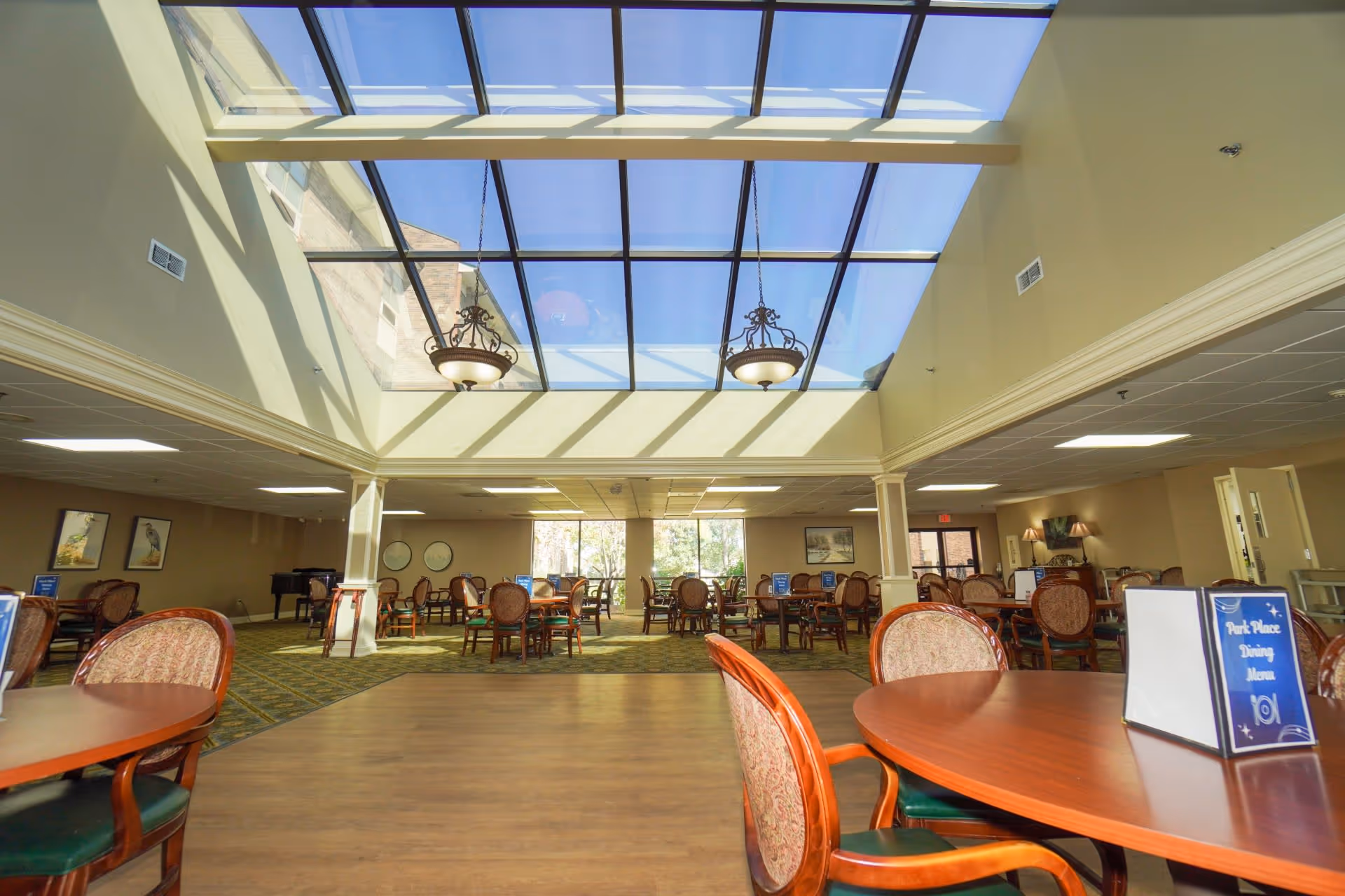 A spacious dining room with multiple round wooden tables and upholstered chairs. The room features a large glass skylight ceiling allowing natural light to fill the space. There are two ornate hanging light fixtures beneath the skylight. The walls are decorated with framed artwork and large windows at the far end provide a view of greenery outside. A sign on one table reads 'Park Place Dining Menu.'