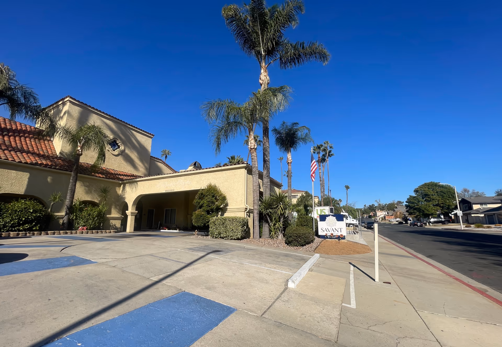Exterior view of Savant of Jurupa Valley facility showing a beige stucco building with a red tile roof, palm trees, bushes, a driveway with blue handicap parking spaces, and a sign with the facility name near the sidewalk under a clear blue sky.