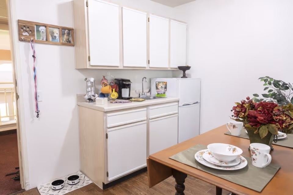 Small kitchenette with white cabinets, a mini fridge and sink, and a dining table set with floral dishes and a vase of flowers.
