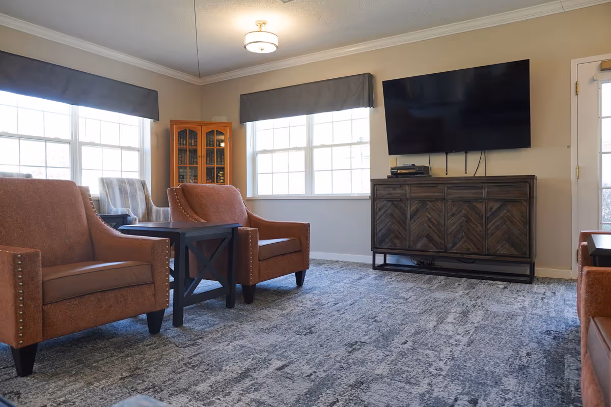 Sunlit communal living room with orange upholstered armchairs, side tables, a wooden media cabinet and a large wall-mounted TV beneath windows.
