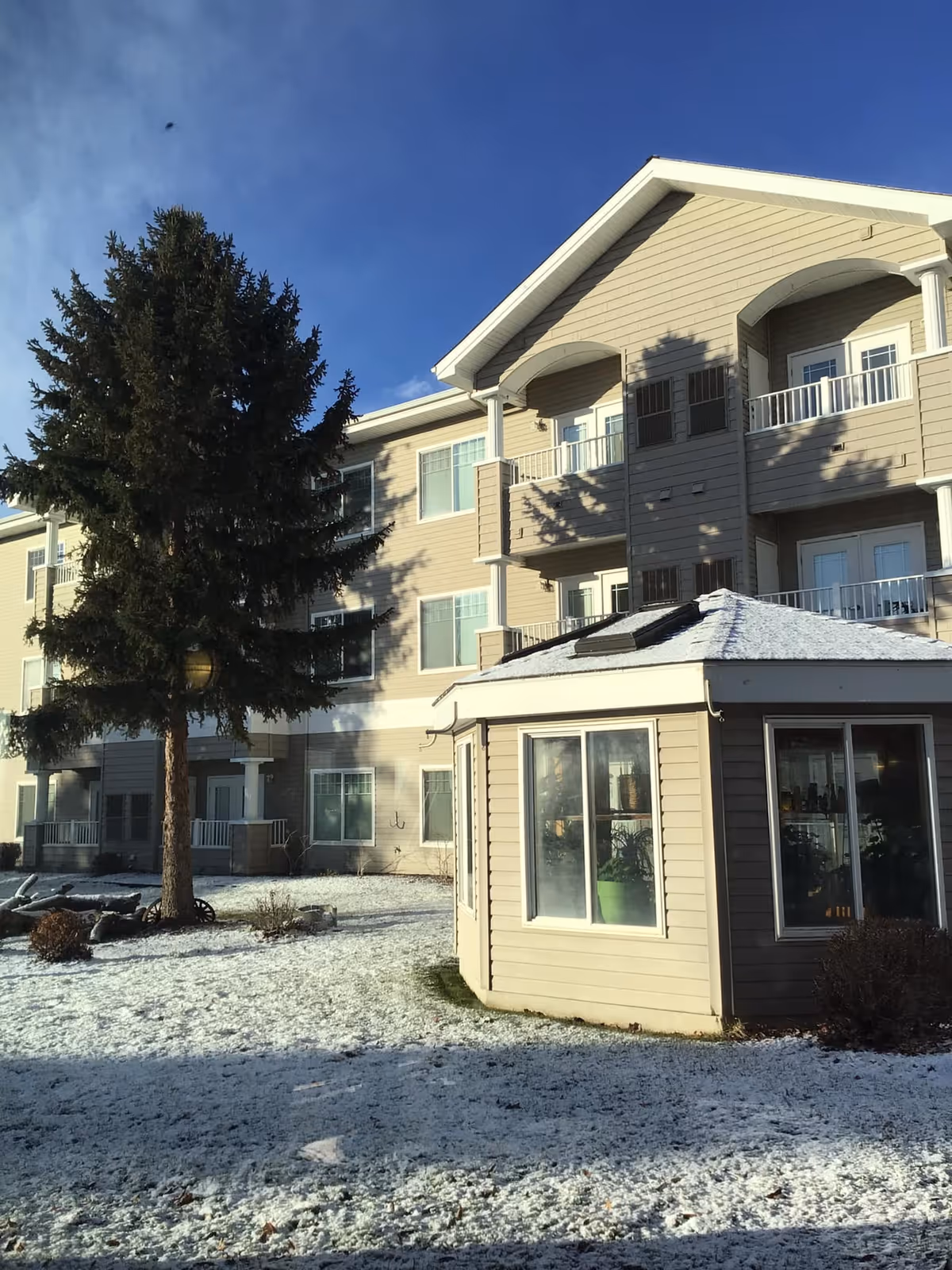 Exterior view of a multi-story residential building with beige siding and white trim under a clear blue sky. In the foreground, there is a small octagonal gazebo with large windows and a snow-covered roof. A tall evergreen tree stands to the left of the gazebo, and the ground is lightly covered with snow.