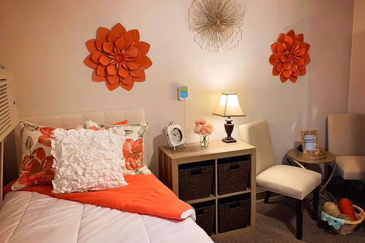 A cozy bedroom corner with a bed featuring floral and white decorative pillows and an orange blanket. Above the bed are two large orange flower wall decorations and one wireframe flower decoration. Next to the bed is a wooden nightstand with a small lamp, a clock, and a vase with pink flowers. Two beige chairs and a small round table with a framed photo and basket with yarn are also visible.