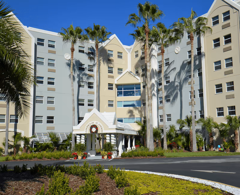 Exterior view of the Landing Of St Andrew senior living facility showing a multi-story building with beige and light blue walls, several tall palm trees, a covered entrance with a wreath, and landscaped greenery in front.