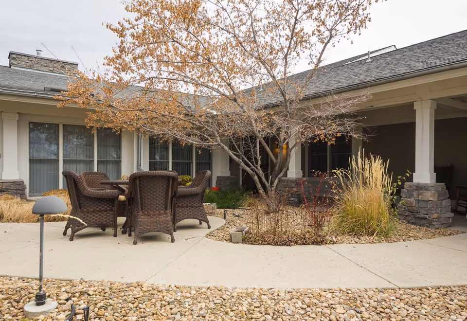 Outdoor courtyard with a round table and wicker chairs beneath a leafless tree in front of a single-story building.