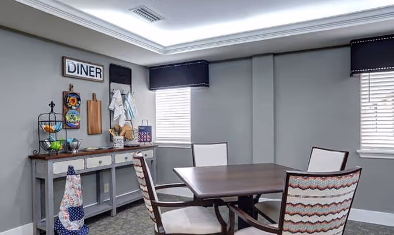 A small dining area with a square wooden table and four upholstered chairs. The walls are painted gray with two windows covered by dark valances. A sideboard against the wall holds kitchen utensils, cutting boards, and decorative items. A sign above the sideboard reads 'DINER'.