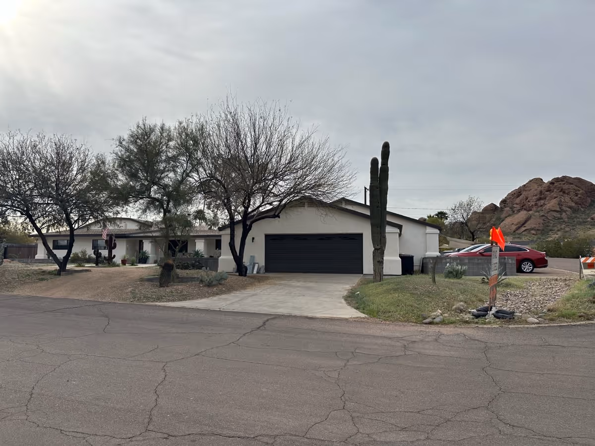 Exterior view of a single-story building with a garage and driveway, surrounded by leafless trees and desert plants, with rocky hills in the background under a cloudy sky.